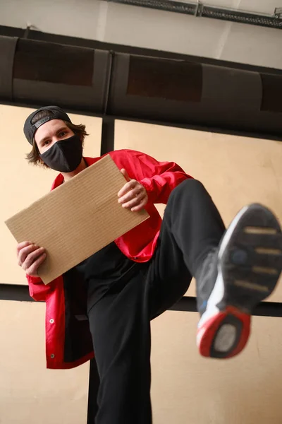 A young man of 25-30 years old in a black protective mask, yellow glasses, a cap and a red jacket holds an empty cardboard sign in his hands.