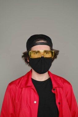 25-30 yaşlarında, siyah koruyucu maskeli, sarı gözlüklü, gri arka planda duran kırmızı ceketli bir genç..
