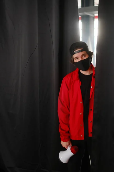 Giovanotto emotivo di 25-30 anni con una maschera protettiva nera, un cappello e una giacca rossa con un megafono su uno sfondo scuro. — Foto Stock