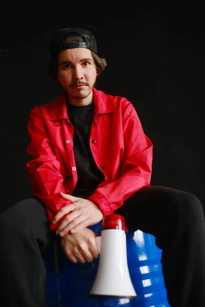 Эмоциональный молодой человек 25-30 лет в черной кепке и в красной куртке сидит на синей бочке и держит в руках мегафон. — стоковое фото