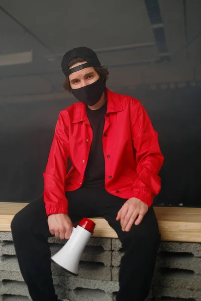 Jeune homme émotionnel de 25 à 30 ans portant un masque de protection noir, une casquette et une veste rouge tenant un mégaphone sur un fond sombre. — Photo