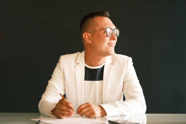 Genç bir öğretmen beyaz ceketli ve gözlüklü bir adam öğrencilerinin ödevlerini kontrol ediyor ve yeni bir derse hazırlanıyor.