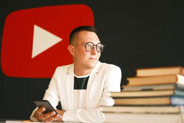 Un hombre de 30-35 años de edad, con un corte de pelo corto, con gafas, una camisa ligera, se sienta en una mesa y se le ocurre un guión para su vlog —  Fotos de Stock