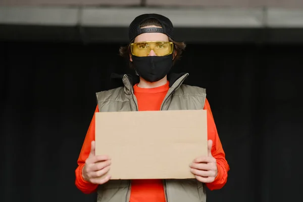 Un jeune homme de 25 à 30 ans portant un masque de protection noir, des lunettes jaunes, une casquette et une veste rouge tient dans ses mains un panneau en carton vide.. — Photo