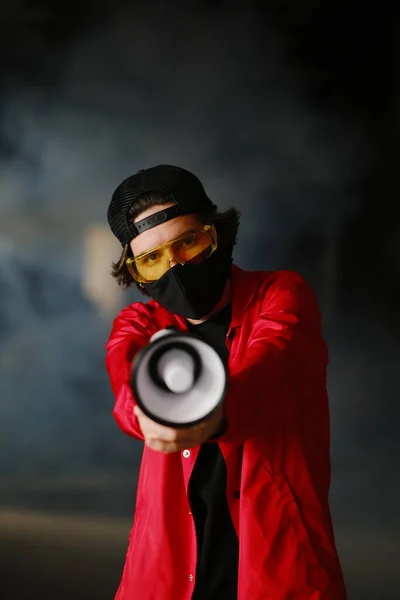 Un jeune homme de 25 à 30 ans portant un masque de protection noir, des lunettes jaunes, un bonnet et une veste rouge tient un mégaphone — Photo