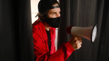 25-30 yaşlarında, siyah koruyucu maskeli, kepli ve kırmızı ceketli koyu bir arka planda megafon tutan duygusal bir genç..
