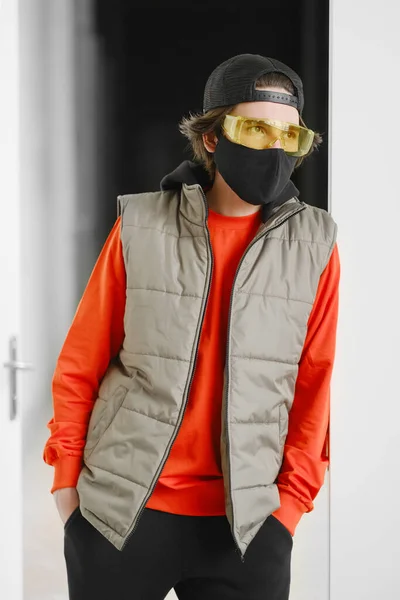 Un joven de 25-30 años con una máscara protectora negra, gafas amarillas, gorra y chaqueta roja posando sobre un fondo gris . — Foto de Stock