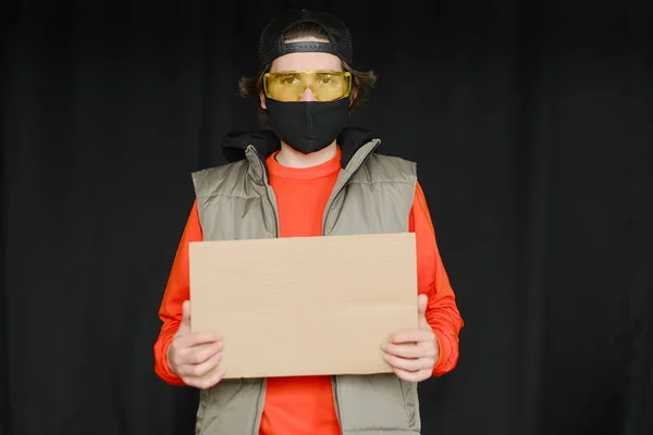 Un jeune homme de 25 à 30 ans portant un masque de protection noir, des lunettes jaunes, une casquette et une veste rouge tient dans ses mains un panneau en carton vide.. — Photo