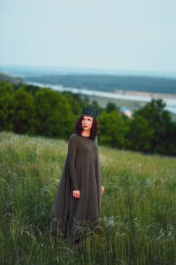 25-30 yaşlarındaki kıvırcık kızıl saçlı, yeşil elbiseli ve tüylü çapraz saçlı genç bir kadın şafak vakti güzel doğanın arka planında duruyor.. 