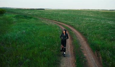 25-30 yaşlarında, kıvırcık kızıl saçlı, siyah deri ceketli ve tüylü bir taç takan genç bir kadın köy yolunda yürüyor..