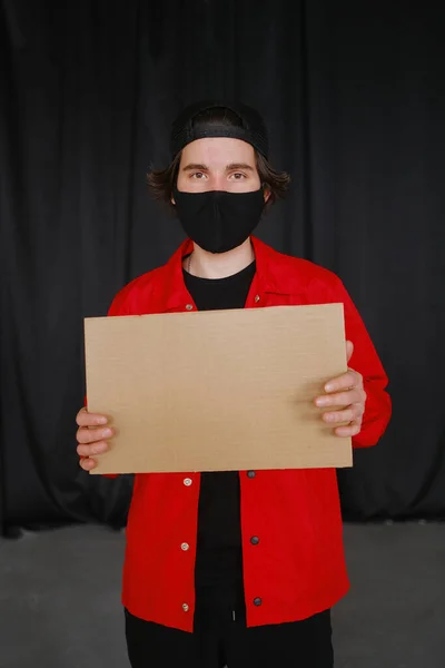 Молодой человек 25-30 лет в черной защитной маске, желтых очках, кепке и красной куртке держит в руках пустой картонный знак. — стоковое фото