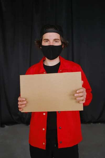 Un joven de 25-30 años con una máscara protectora negra, gafas amarillas, una gorra y una chaqueta roja sostiene un cartel de cartón vacío en sus manos . — Foto de Stock