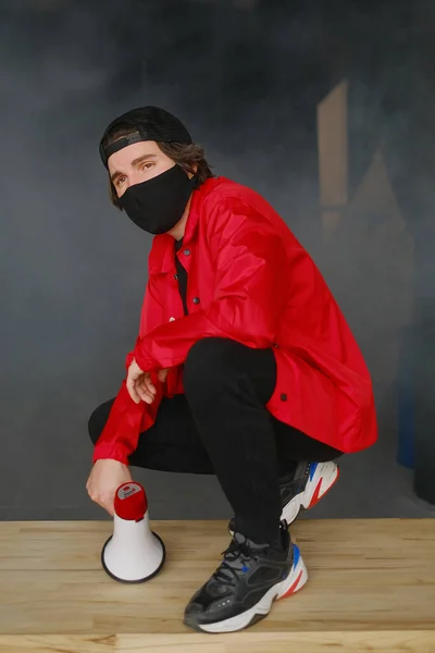 Эмоциональный молодой человек 25-30 лет в черной защитной маске, кепке и красной куртке, держащий мегафон на темном фоне. — стоковое фото