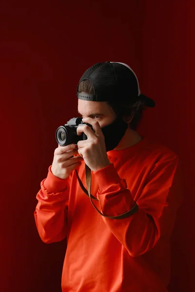 Photographe masculin. Un jeune homme de 25 à 30 ans portant un masque de protection noir et un sweat-shirt rouge tient dans ses mains un appareil photo sans miroir. — Photo