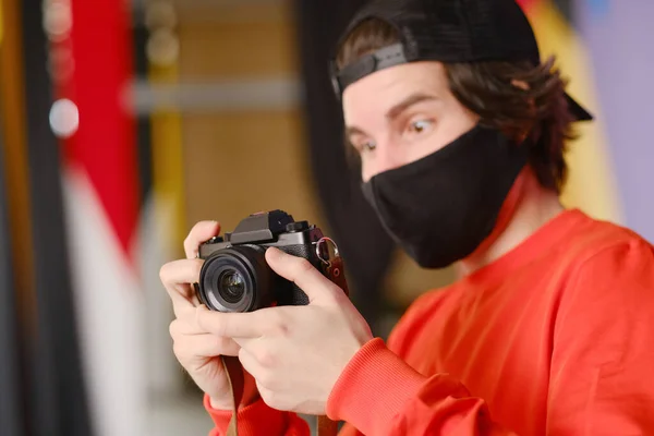 Fotógrafo masculino. Un joven de 25-30 años con una máscara protectora negra y una sudadera roja sostiene una cámara sin espejo en sus manos . Fotos De Stock