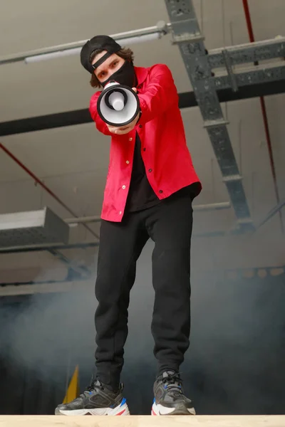 Jeune homme émotionnel de 25 à 30 ans portant un masque de protection noir, une casquette et une veste rouge tenant un mégaphone sur un fond sombre. Photo De Stock