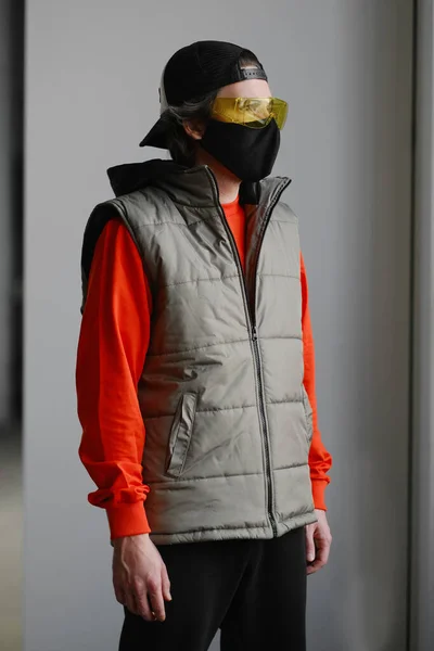 Un jeune homme de 25 à 30 ans portant un masque de protection noir, des lunettes jaunes, un bonnet et une veste rouge posant sur un fond gris. — Photo