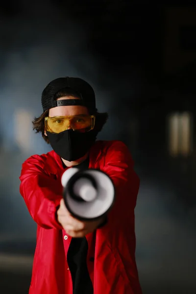 Un giovane di 25-30 anni con una maschera protettiva nera, occhiali gialli, un cappello e una giacca rossa regge un megafono — Foto Stock