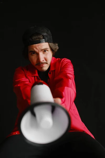 Un jeune homme émotionnel de 25 à 30 ans coiffé d'une casquette noire et d'une veste rouge est assis sur un tonneau bleu et tient un mégaphone dans ses mains sur un fond sombre. Concept de protestation Photo De Stock