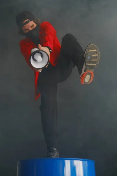 Un joven emocional de 25-30 años con una máscara protectora negra, una gorra y una chaqueta roja sostiene un megáfono sobre un fondo oscuro . Imágenes De Stock Sin Royalties Gratis