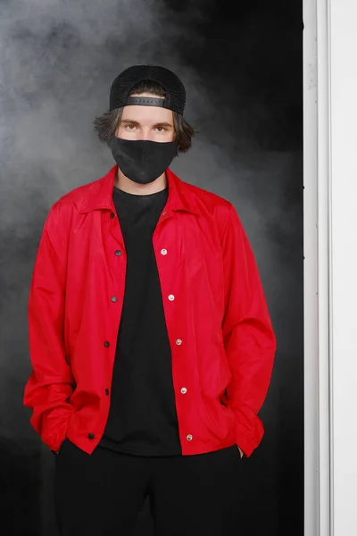 Portrait d'un jeune homme de 25 à 30 ans portant un masque de protection noir, une casquette et une veste rouge. Image En Vente