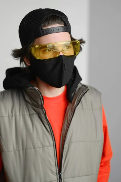 Un joven de 25-30 años con una máscara protectora negra, gafas amarillas, gorra y chaqueta roja posando sobre un fondo gris . Fotos De Stock Sin Royalties Gratis