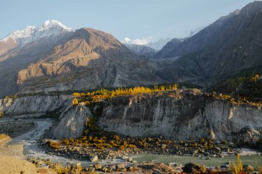 Karakoram sıradağlarındaki karla kaplı dağ zirvelerine karşı renkli ağaçlarda ve nehirde parlayan doğal güneş ışığı. Hunza Nagar Vadisi sonbahar mevsiminde. Gilgit Baltistan, Pakistan.