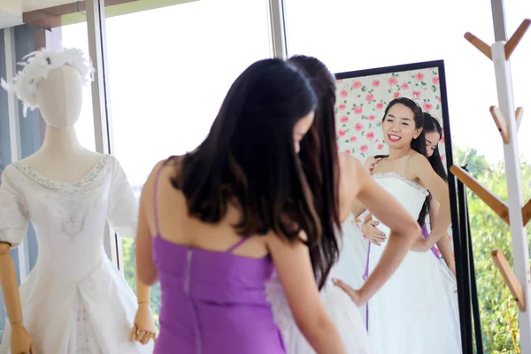 Beautiful bride getting dressed by her best friend in her wedding day and choosing a wedding dress in the shop and the shop assistant is helping her