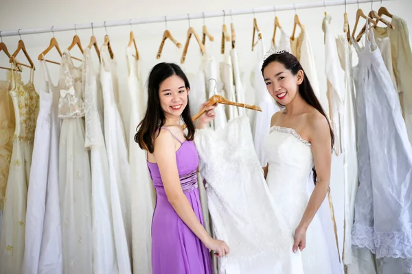 Beautiful bride getting dressed by her best friend in her wedding day and choosing a wedding dress in the shop and the shop assistant is helping her