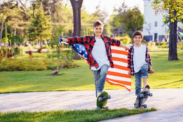 America\'s USA Independence Day. Two cute brothers guy americans standing in a city park on skateboards and holding flag of USA. Stretching fabric and shouting exiting with joy in flannel shirts.