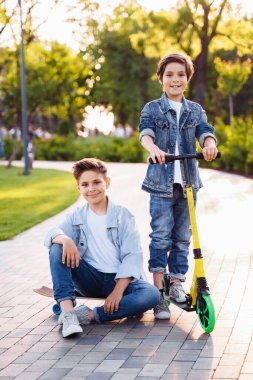 Yaz parkında aktif çocuk eğlencesi. Bir yaz akşamında parkta eğlenen iki sevimli kardeş. Kaykay ve scooter 'a binmek. Kot pantolon ve beyaz tişörtler giymişti..