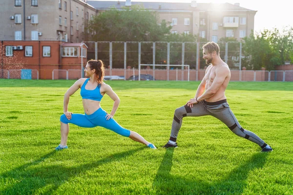 魅力的な若いカップルの男性と女性25歳は 芝生のスポーツ競技場で晴れた夏の朝に演習を行っています 喜びと笑顔で斜面を作り 筋肉を伸ばし 足を上げる — ストック写真