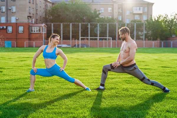 魅力的な若いカップルの男性と女性25歳は 芝生のスポーツ競技場で晴れた夏の朝に演習を行っています 喜びと笑顔で斜面を作り 筋肉を伸ばし 足を上げる — ストック写真