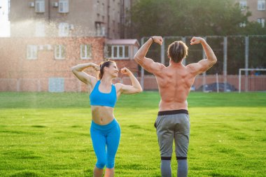 İkişerli egzersizler. Çekici sporcular kız ve erkek pompalanmış vücutlarını gösteriyorlar. Adam çıplak bir gövdeyle duruyor, kız mavi bir bluz ve tayt giyiyor. Kollardaki kasları gösteriyor..