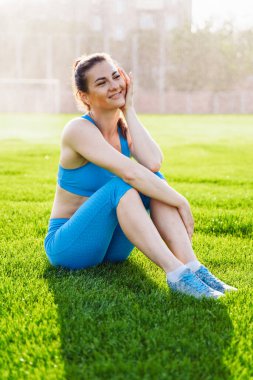 Güzel, kaslı bir vücudu olan atletik bir kadın şehir futbol stadyumunda çimlerin üzerinde oturuyor. Sıcak bir yaz akşamında, arka planda akan suyun gülümsemesi..