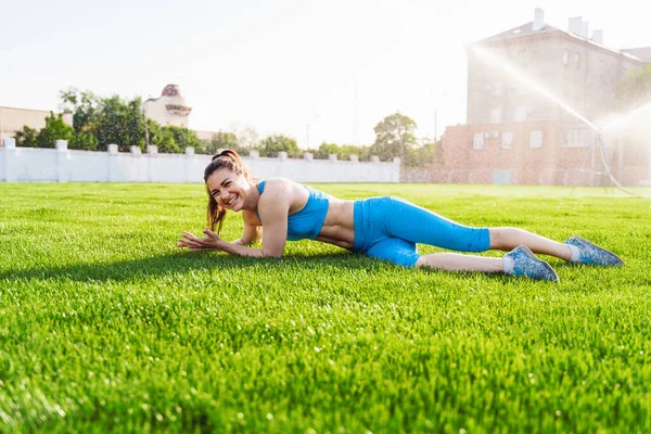 芝生のスタジアムに横たわって青いフィットネス服の女の子 ブルネットはサッカー場の緑の芝生を宣伝している 女性はスポーティーなライフスタイル シティスタジアムでのトレーニング後に休憩 ポーズモデル — ストック写真