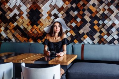 Sabahın erken saatlerinde şirin ve şık bir kafede. Siyah, minimalist bir elbise giymiş genç bir kadın elinde siyah bir fincan kahve ya da çayla ahşap bir masada oturuyor. Sakin bir gün, dinlendirici bir ortam..