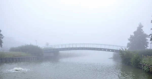 On a thick foggy morning along the water there is a foot or bike bridge