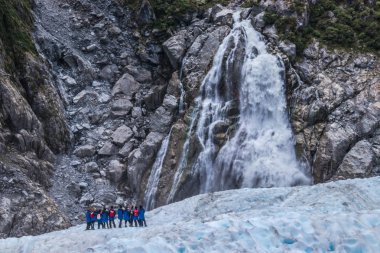 Turist üzerinde vahşi karlı arazi kayalık uçurum ile su cascade, South Island altında mesafede Grup