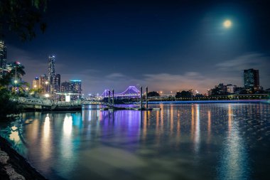 Sahil Manzaralı parlayan ile modern ışıklı şehir mor Köprüsü ve kalın yemyeşil ağaçlar pier üzerine ay ışığı altında Nehri'nin sakin su