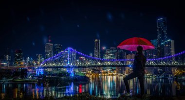 Ayakta ve gece Brisbane ışıklı köprü ve gökdelenler ile modern şehir manzaralı tepede kırmızı şemsiye tutan anonim adam