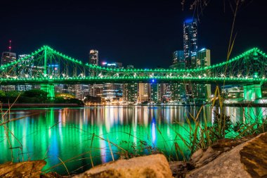 Rock ve çim kenarı parlak parlayan Köprüsü ve nehrin karşı kıyısında gece zamanında bina bitti