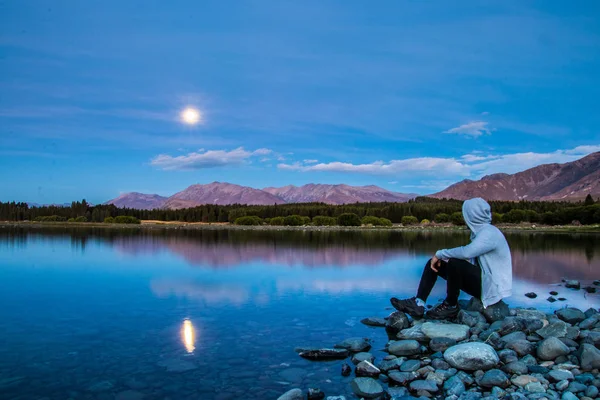 Vista Lateral Homem Sentado Costa Rochosa Com Água Reflexão Montanhas Fotos De Bancos De Imagens