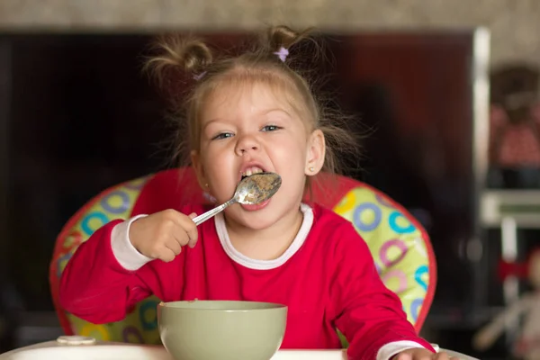 Retrato de menina lambendo colher comer mingau sentado na cadeira de alimentação — Fotografia de Stock