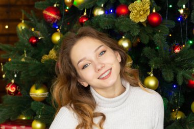 Sandalyede oturan ve Noel ağacı arkaplanındaki kameraya bakan gülümseyen genç kadının portresi.