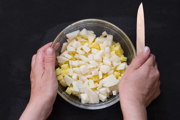 Vue du dessus de la pomme de terre crue coupée dans un bol en verre et les mains de la femme avec un couteau sur le fond noir — Photo