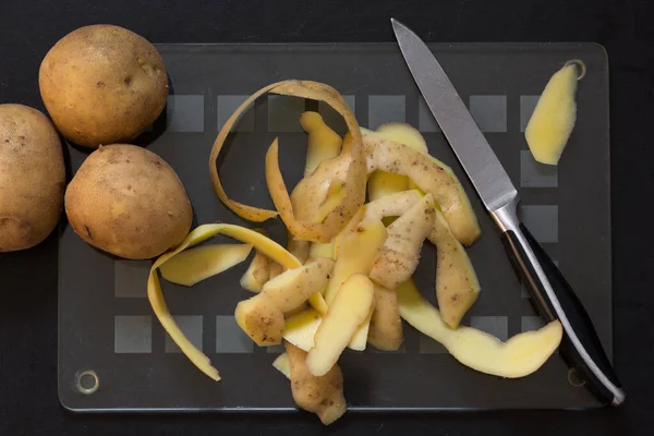 Draufsicht auf rohe, ungeschälte Kartoffeln mit Schalen und das normale Leben auf einem Glasschneidebrett auf schwarzem Hintergrund — Stockfoto