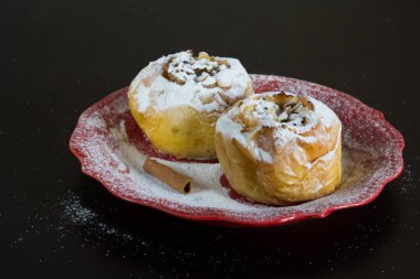 İki fırında elma yulaf dolu ve siyah zemin üzerinde kırmızı tabakta şeker tozu ile pudralanmış.