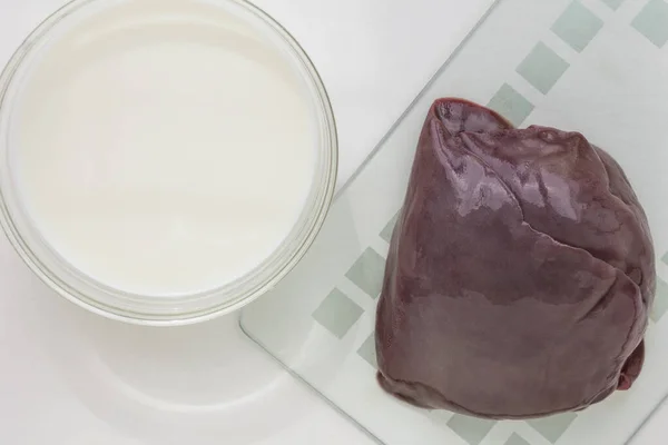 Вид сверху сырого куска печени на стеклянной тарелке и молока в миске на белом фоне — стоковое фото