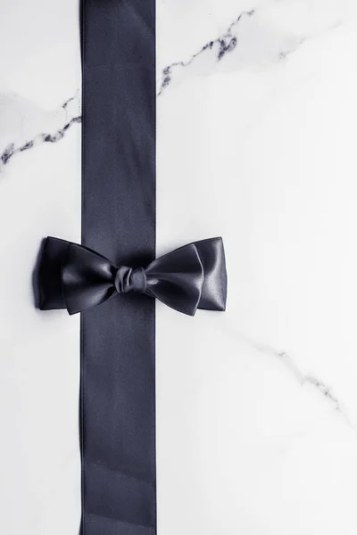 Black silk ribbon and bow on marble background, flatlay